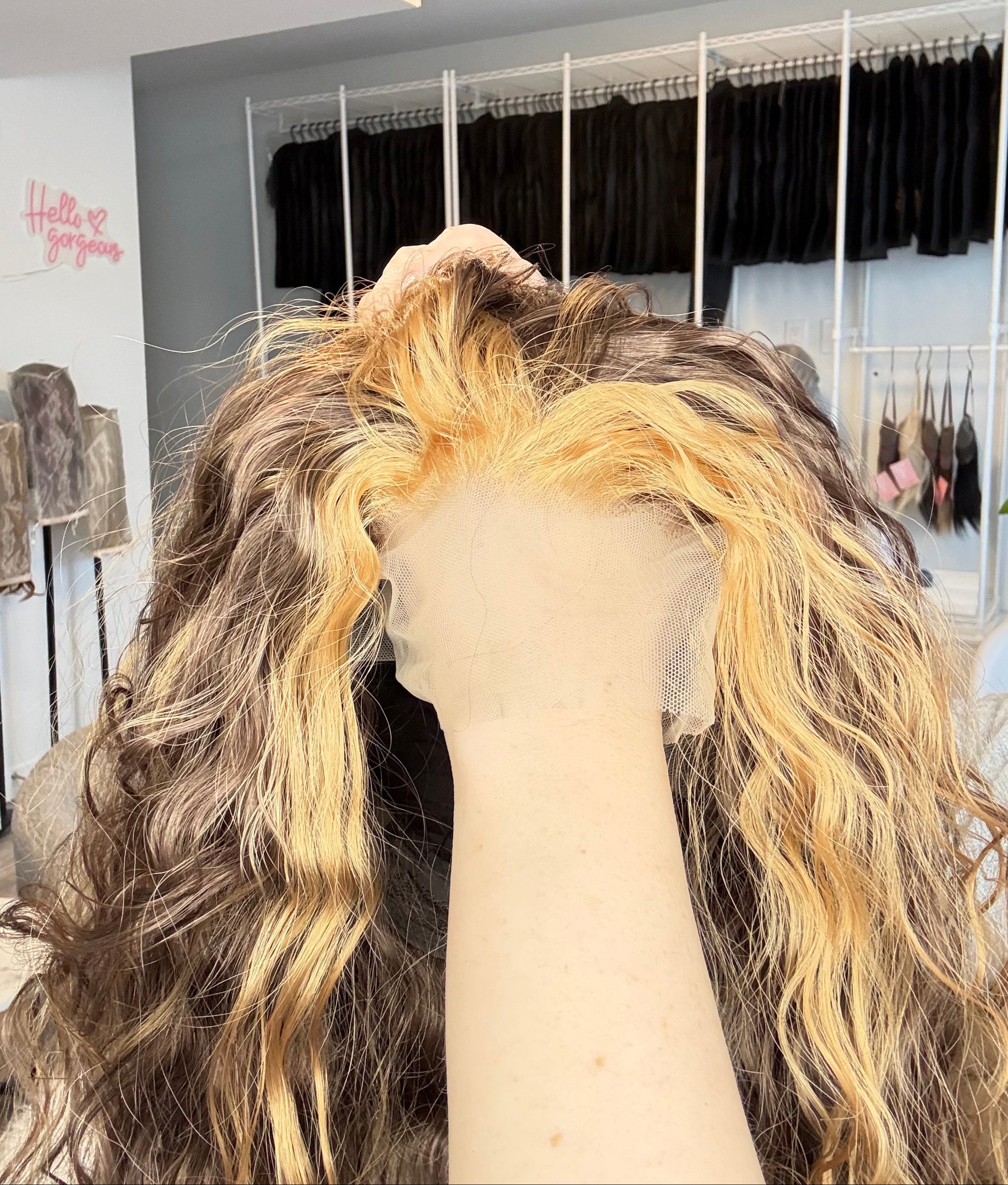 Close-up of a wig with blonde and brown hair on a mannequin head, with a mirror and light fixture in the background.