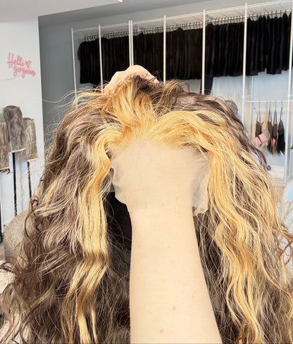 Close-up of a wig with blonde and brown hair on a mannequin head, with a mirror and light fixture in the background.