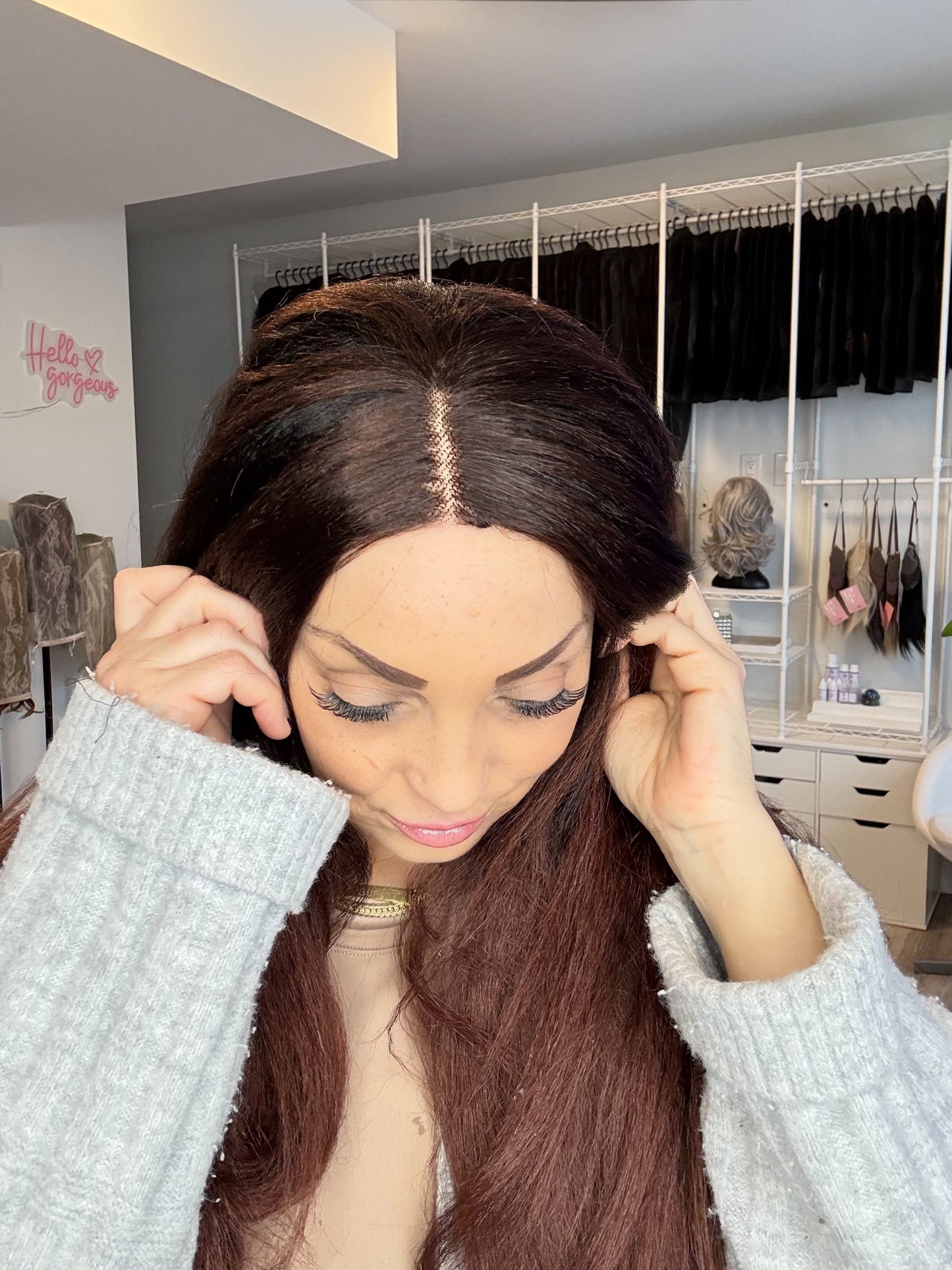 Woman adjusting a brown wig in a room with clothing racks.
