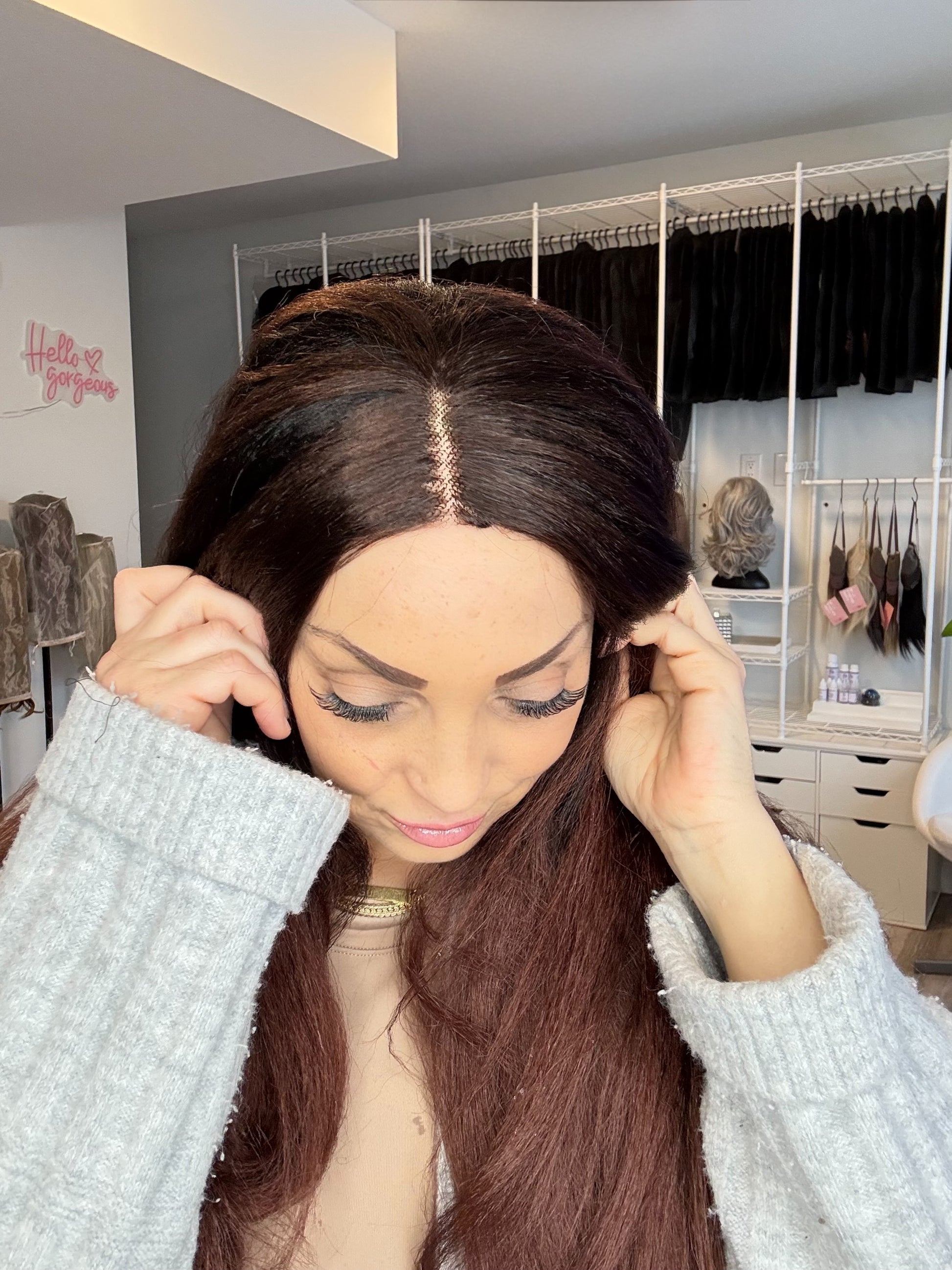 Woman adjusting a brown wig in a room with clothing racks.
