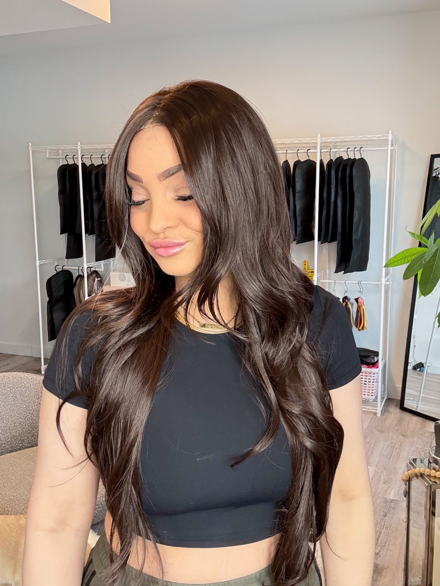 Woman with long, wavy hair wearing a black crop top and green pants in a room with clothing racks and plants.
