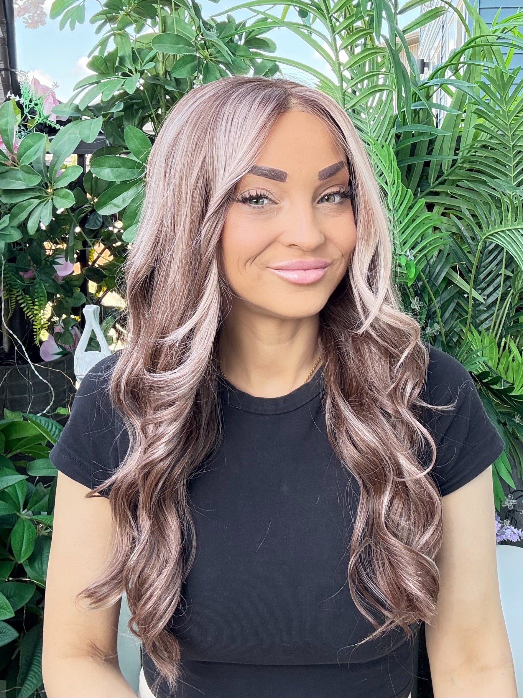 Woman with long, wavy hair standing outdoors with greenery in the background