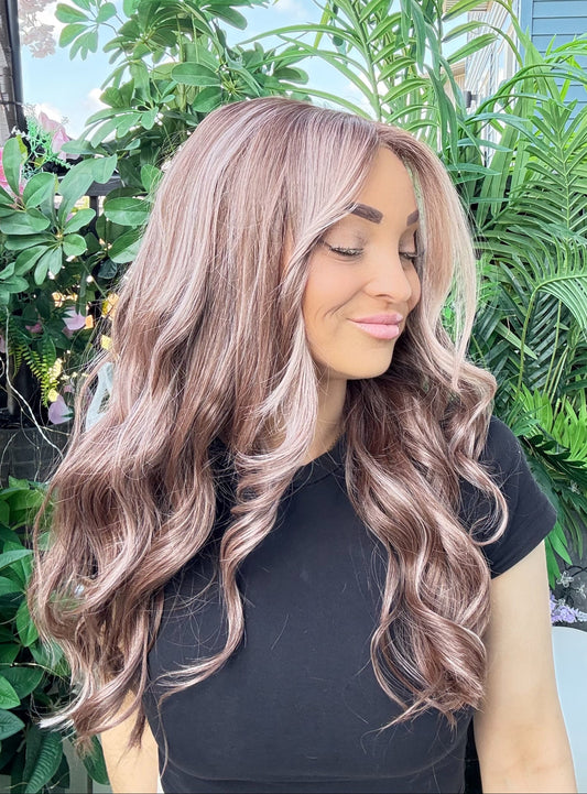 Woman with long, wavy, pinkish-brown hair standing outdoors with greenery in the background
