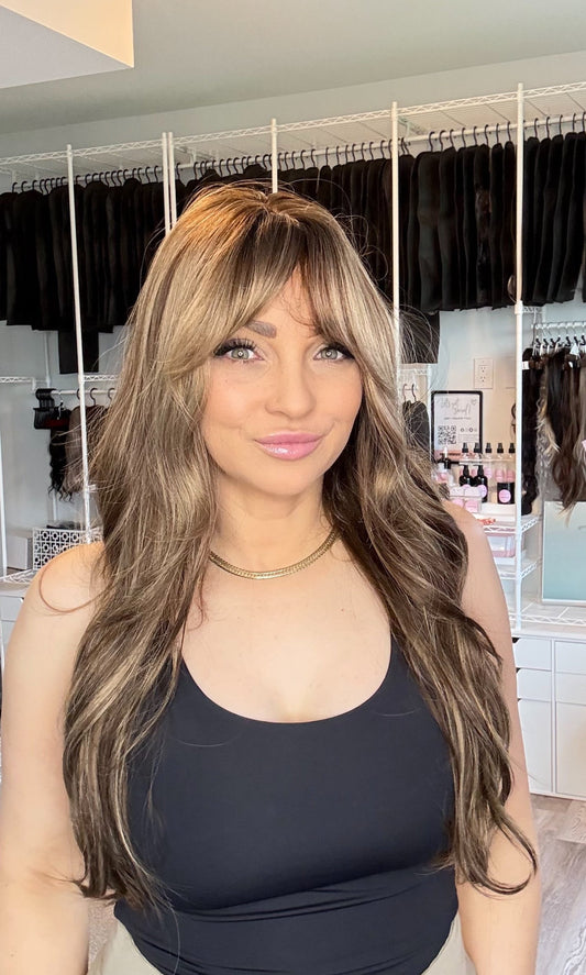A woman modeling a long, wavy wig with light brown and blonde highlights, featuring a straight-cut bang, in a hair salon setting.
