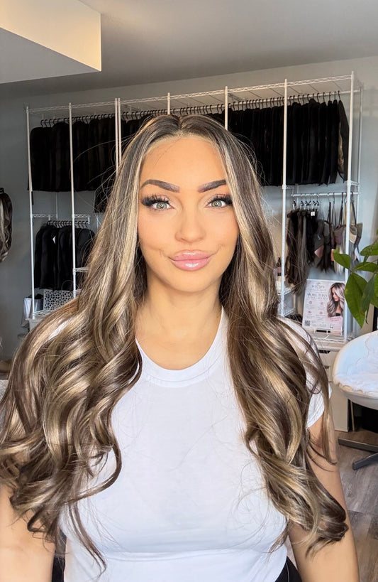 A woman with long, wavy, highlighted hair wearing a white t-shirt, standing in front of a hair salon closet rack.