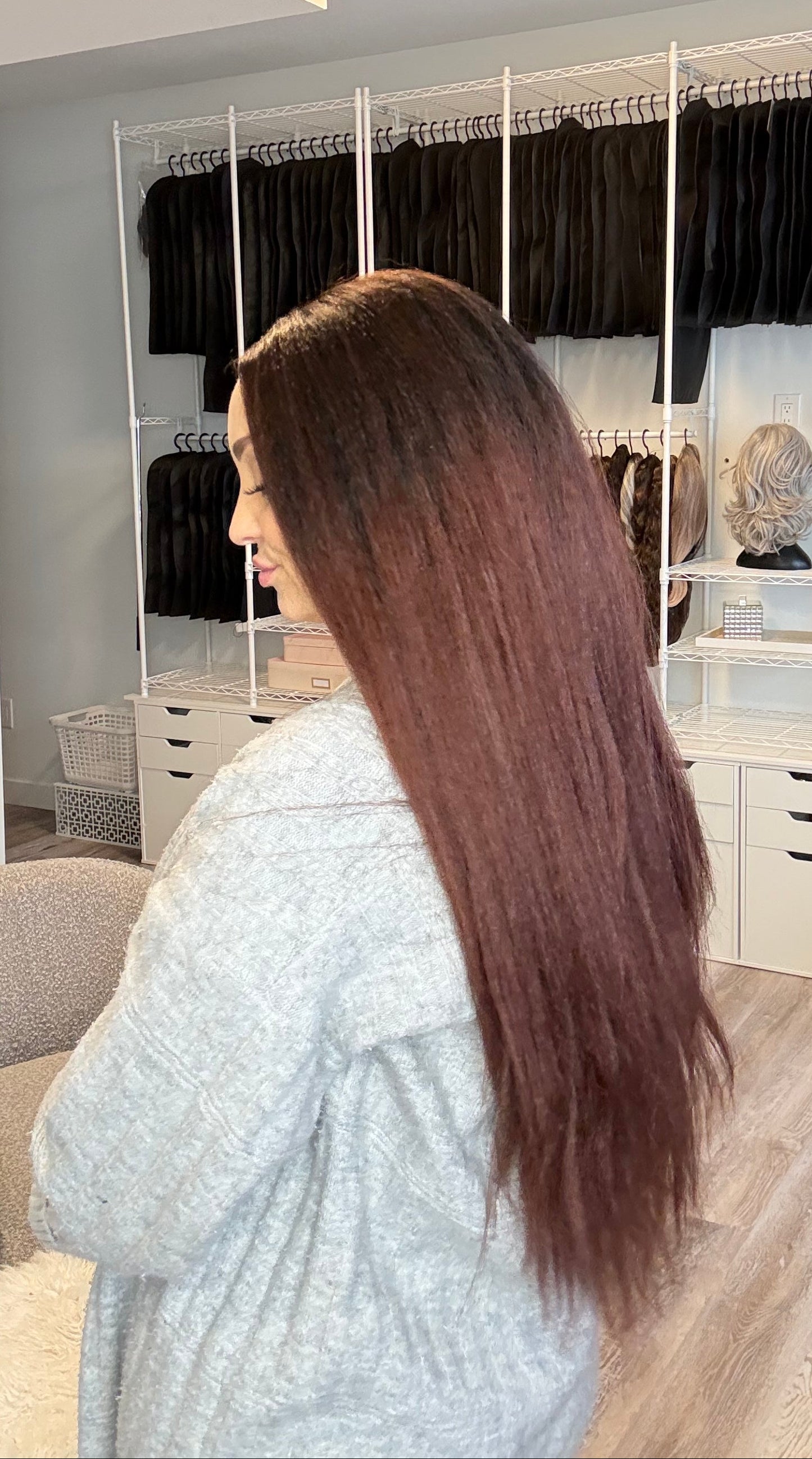 Woman with long hair in a salon setting, looking at hair extensions displayed on a rack.