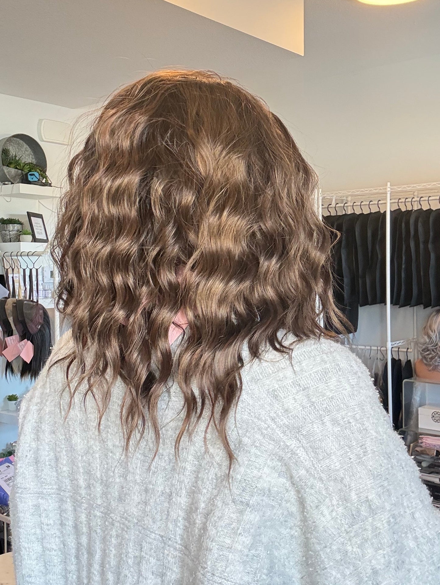 Person with wavy hair standing in a room with a mirror and various items on shelves.