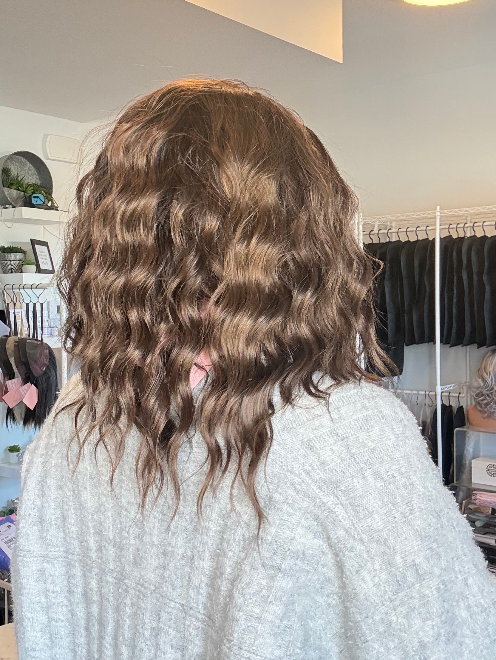 Person with wavy hair standing in a room with a mirror and various items on shelves.