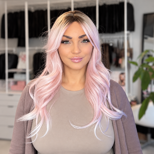 A woman with long, wavy pastel pink and white ombre hair wearing a taupe top and cardigan, standing in front of a blurred closet background.