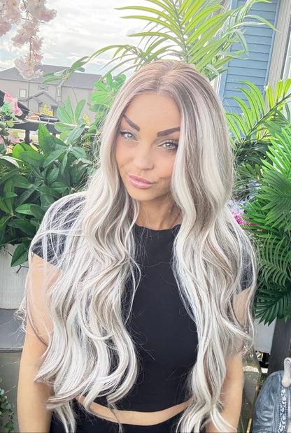 Woman with long, wavy, platinum blonde hair standing outdoors with greenery in the background