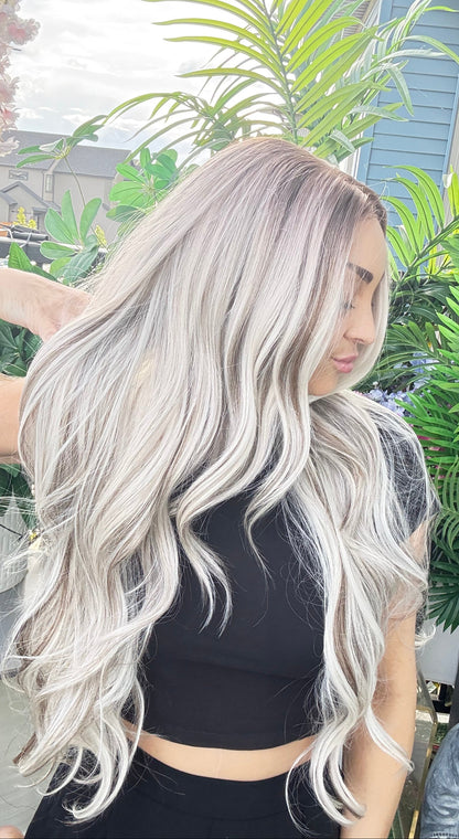Person with long, wavy, silver hair standing among green plants.