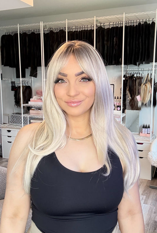 A woman with long platinum blonde hair and bangs wearing a black tank top, standing in front of a white clothing rack filled with black garments in a room.