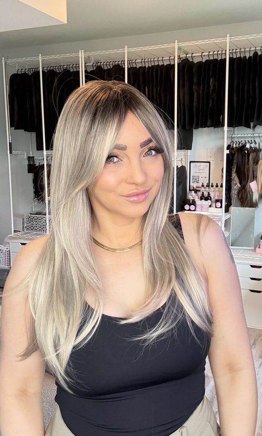 A woman with a long, straight blonde wig with dark roots is wearing a black tank top and beige pants, standing in a room with a white clothing rack in the background.