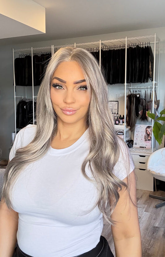 A woman with long, wavy silver-gray hair wearing a white t-shirt, standing in front of a closet rack filled with black clothing.