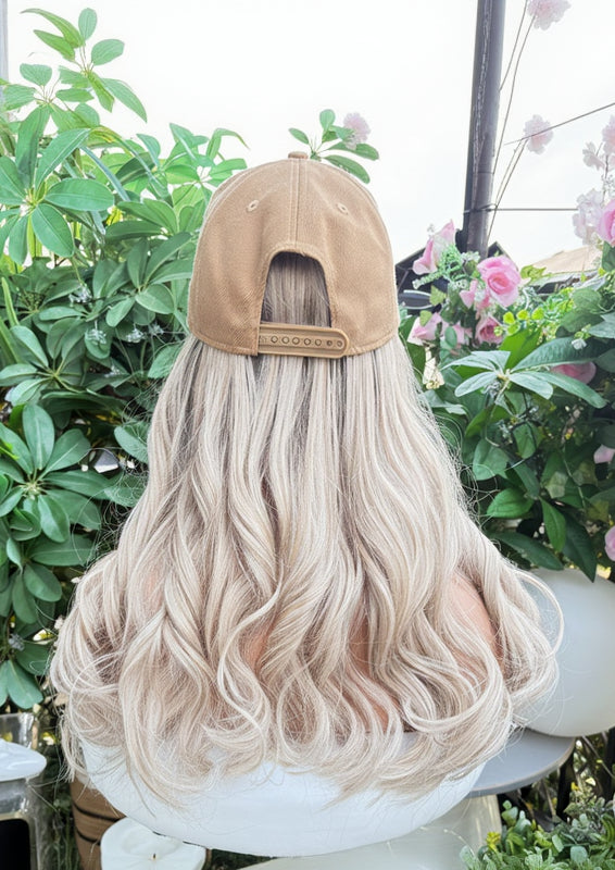 Blonde wig on a mannequin head with a beige cap, set against a decorative background with plants and flowers.