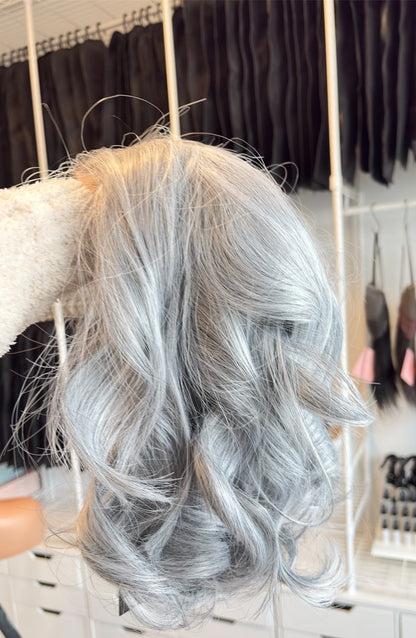 Gray wig held by a person with a blurred background of hair extensions and tools.
