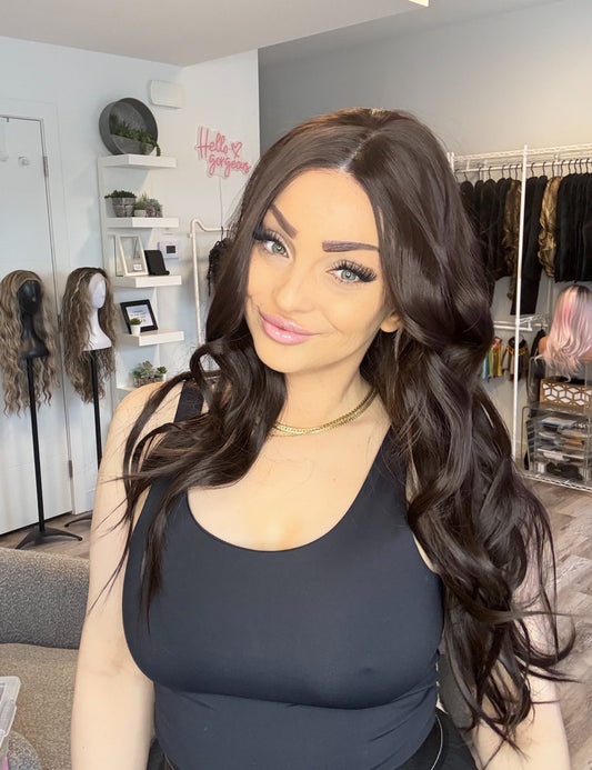 Woman with long dark hair in a salon setting with wigs and styling tools visible.