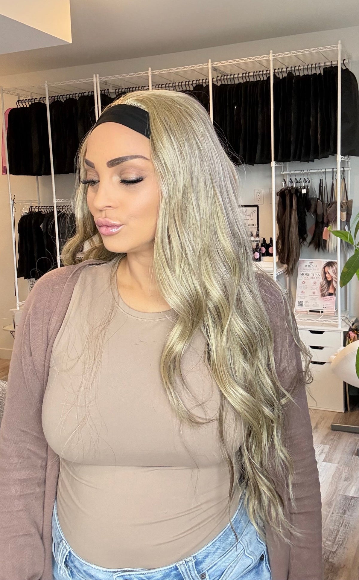 A woman with long, wavy platinum blonde hair wearing a black headband, beige bodysuit, and brown cardigan, standing in a salon or dressing room with clothing racks and a plant in the background.
