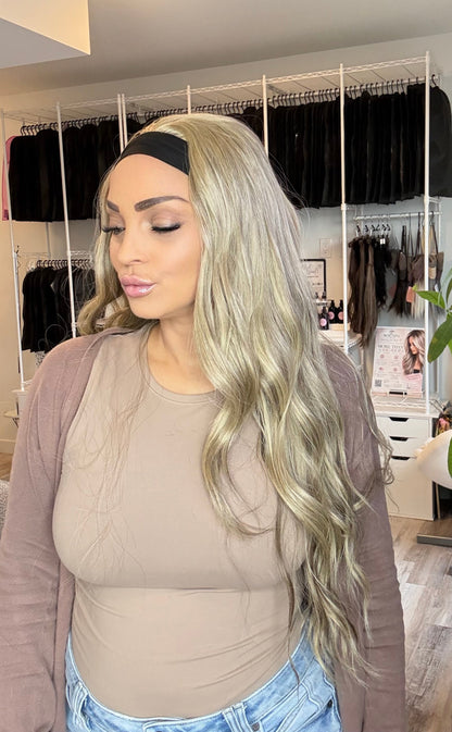 A woman with long, wavy platinum blonde hair wearing a black headband, beige bodysuit, and brown cardigan, standing in a salon or dressing room with clothing racks and a plant in the background.