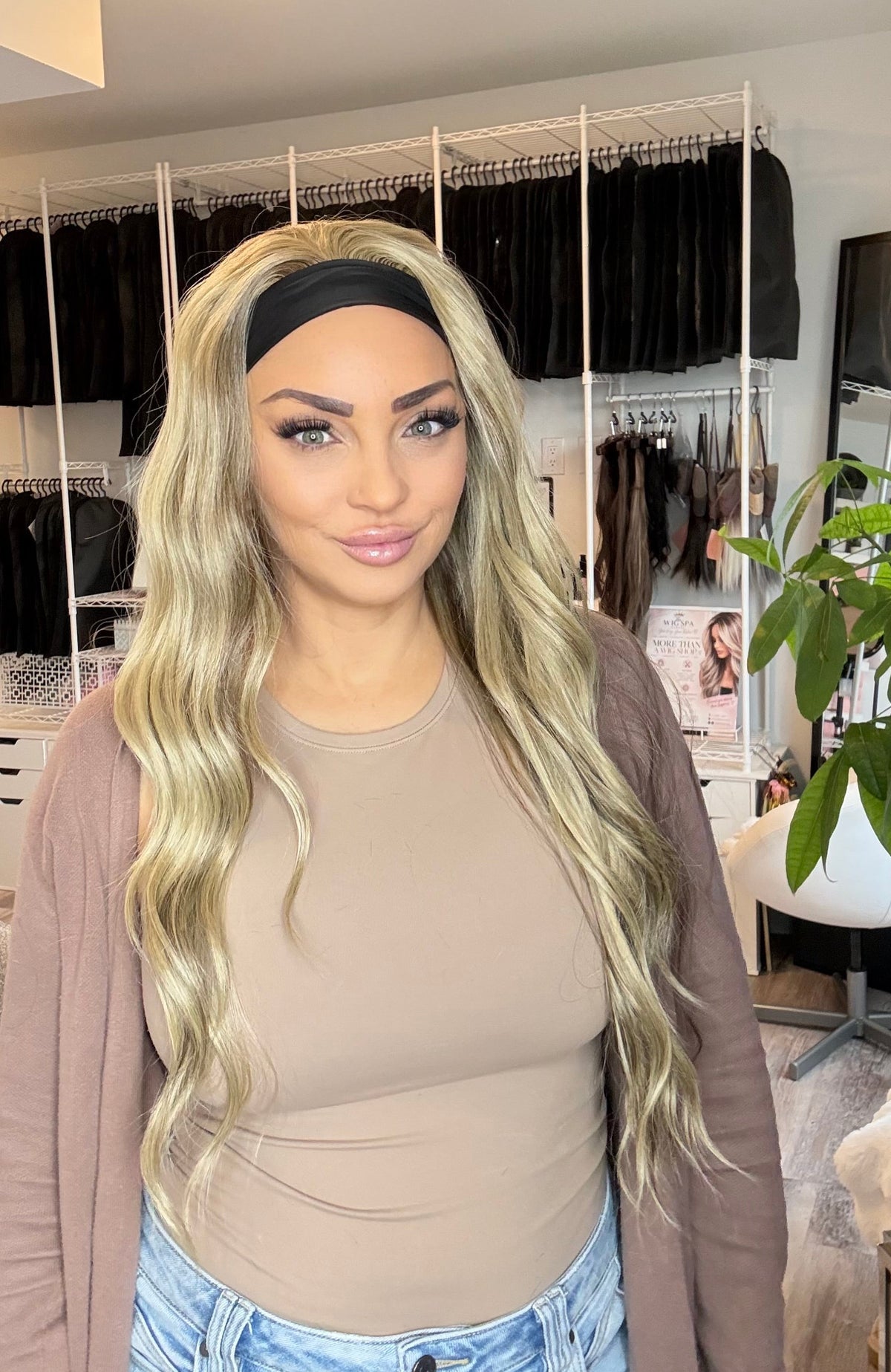 A woman with long, wavy blonde hair wearing a black headband, a beige ribbed top, and a brown cardigan, standing in a room with a clothing rack and plant in the background.
