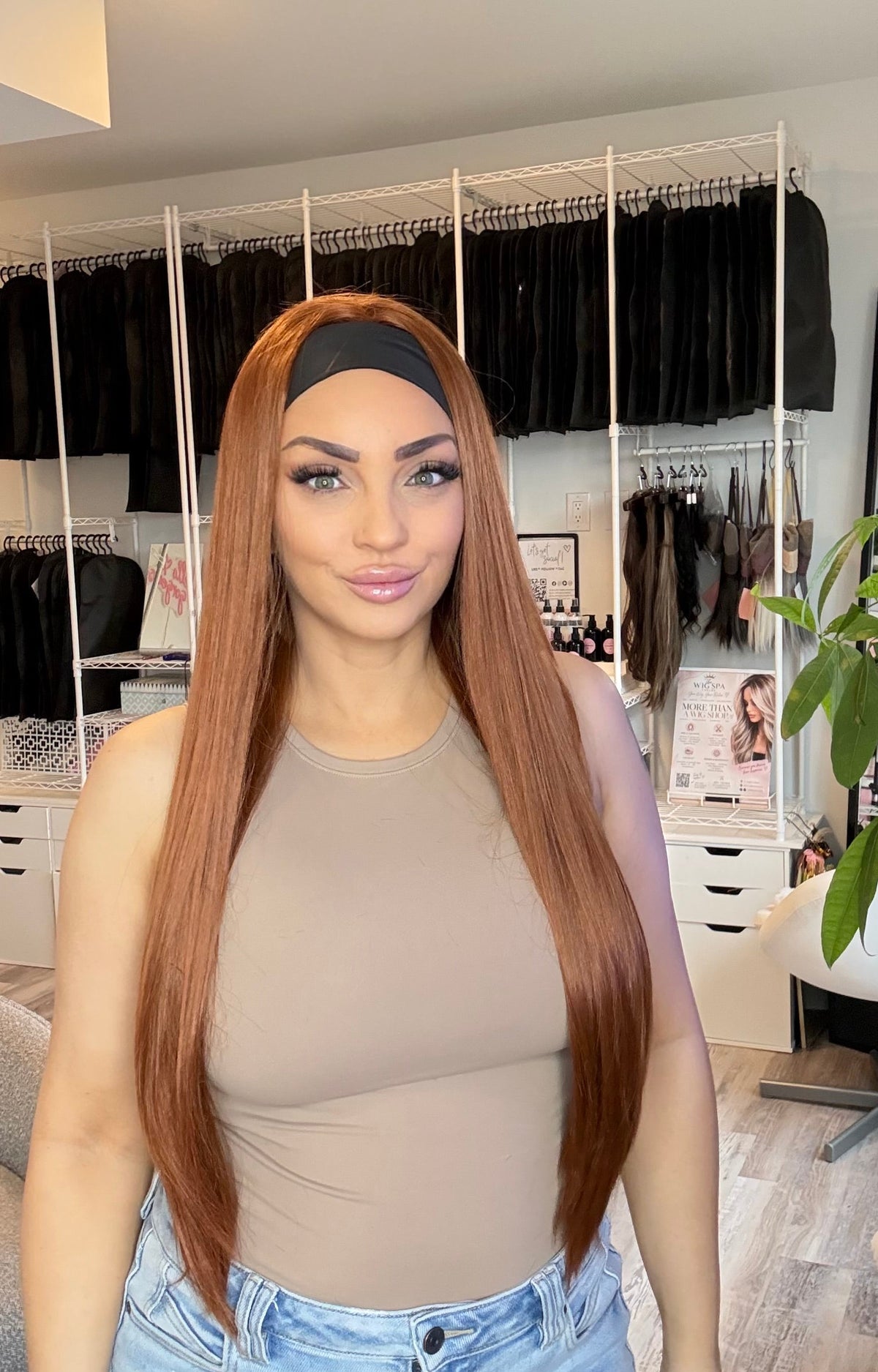 A woman with long, straight, auburn-brown hair wearing a black headband and a beige top, standing in a room with clothing racks and a plant in the background.