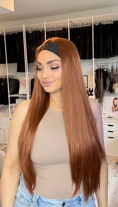 A woman with long, straight, auburn-brown hair wearing a black headband and a beige top, standing in a modern room with a clothing rack and plant in the background.