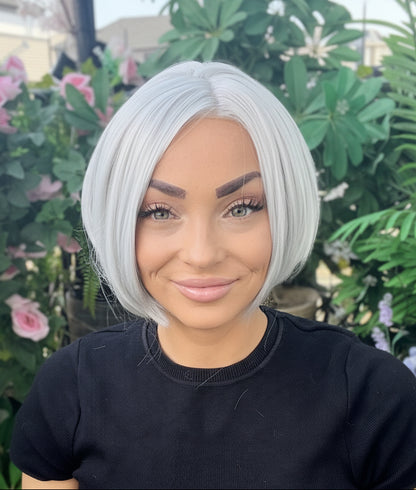 Person with a short, styled haircut standing outdoors with greenery and flowers in the background