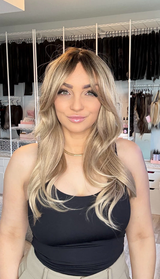 A woman with long blonde-highlighted wavy hair wearing a black tank top and beige pants, standing in front of a white clothing rack filled with black garments in a room.