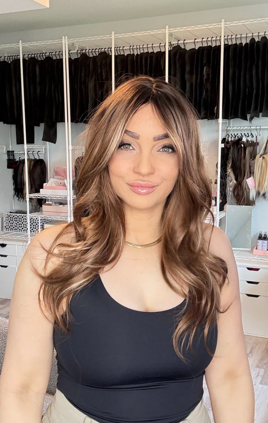 A woman with long, wavy brown hair wearing a black tank top and beige pants, standing in a room with a large clothing rack in the background.