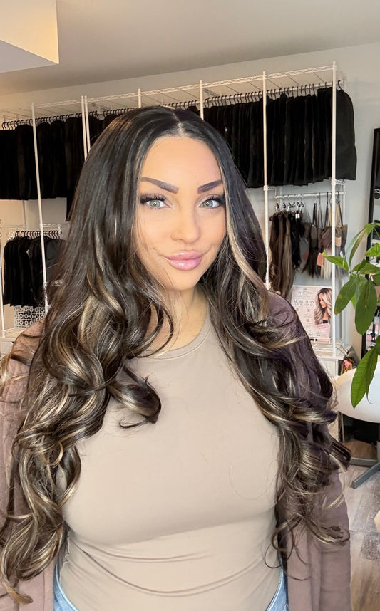 A woman with long, wavy, dark brown hair with lighter brown highlights is wearing a beige top and a brown jacket, standing in a room with a clothing rack and a plant in the background.