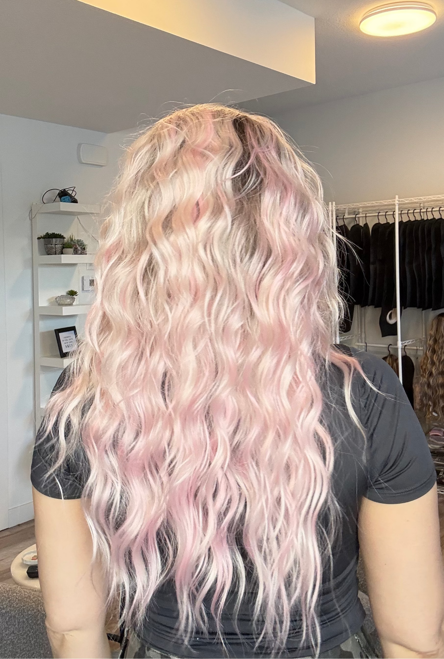 Person with long, wavy, pink hair in a room with a mirror and clothing rack.
