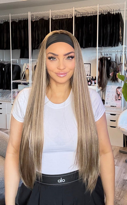 A woman with long, straight, light brown to blonde highlighted hair is shown indoors, wearing a white t-shirt and a black skirt. She is standing in front of a clothing rack filled with black wigs or hairpieces in a room with a salon-like interior.