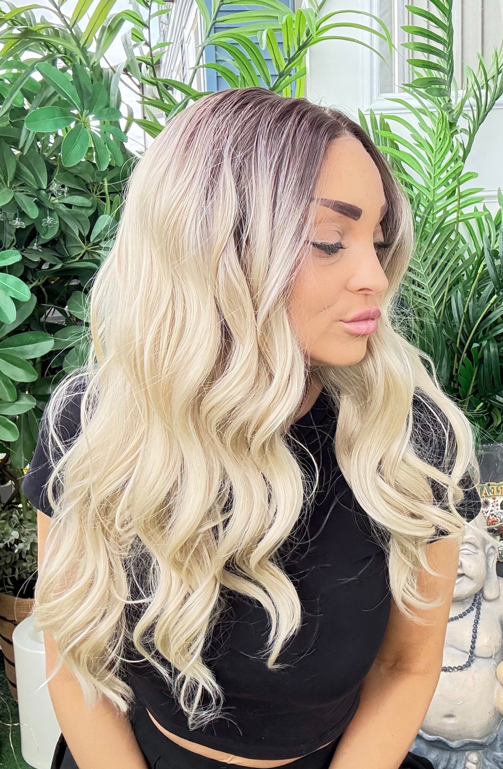 Woman with long, wavy blonde hair sitting outdoors with greenery in the background