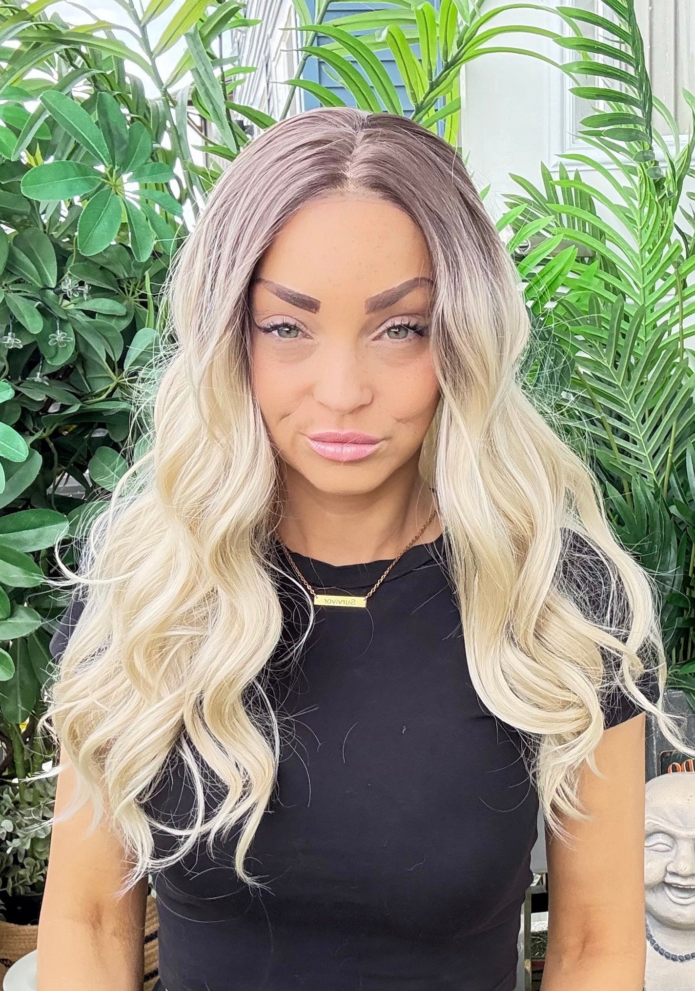 Person with long blonde hair in a black outfit standing in front of green plants.