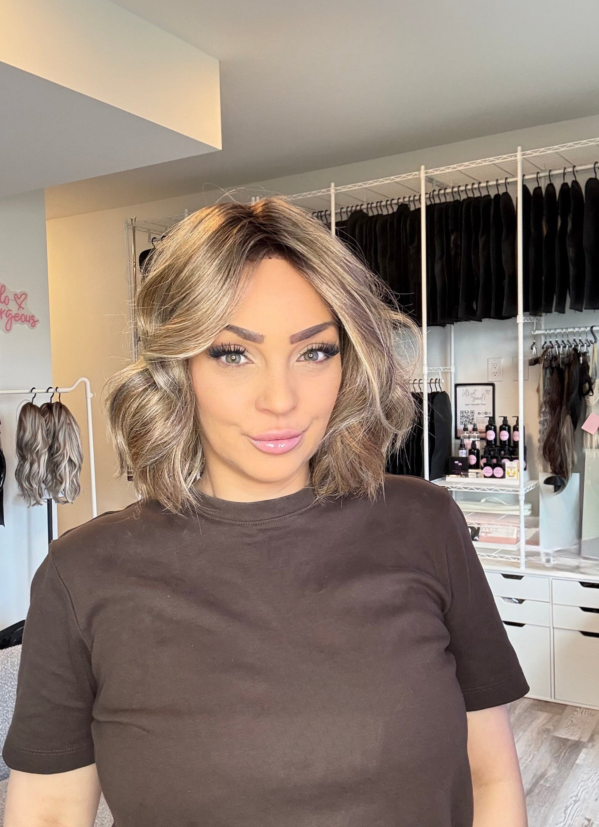 A woman with shoulder-length blonde-highlighted wavy hair wearing a dark brown t-shirt, standing in a room with a closet background.
