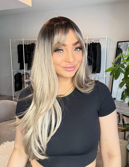 Woman with long blonde hair in a room with clothing racks and plants.