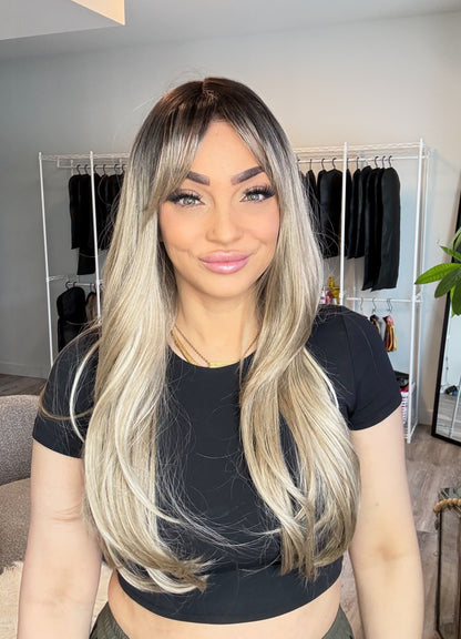 Woman with long blonde hair in a room with clothing racks and a plant.
