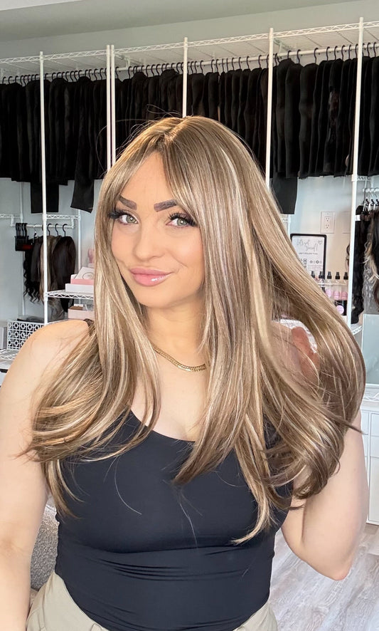 A woman with a long, wavy blonde wig is wearing a black tank top and beige pants in a room with a clothing rack in the background.
