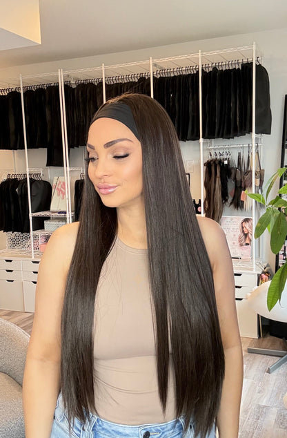 A woman with very long, straight, dark brown to black hair is shown indoors, wearing a beige ribbed tank top and light blue jeans. She is standing in front of a white clothing rack filled with black garments in a room with light wood floors and a green plant.