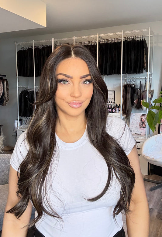 A woman with long, wavy, dark brown hair with subtle lighter highlights, wearing a white t-shirt in a boutique setting with clothing racks and a plant in the background.
