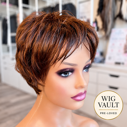A short, layered wig with warm medium brown and caramel highlights, displayed on a mannequin head in a boutique setting. A circular 'WIG VAULT PRE-LOVED' badge is visible in the bottom right corner.