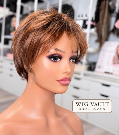 A short, layered wig with brown and blonde highlights displayed on a mannequin head in a boutique setting. A 'WIG VAULT PRE-LOVED' label is visible in the bottom right corner.